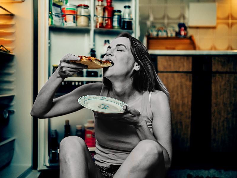 Woman eating pizza slice in front of the refrigerator